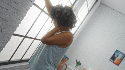 Young woman feeling sick leaning against the wall in a bright bedroom with curly hair and a blue...