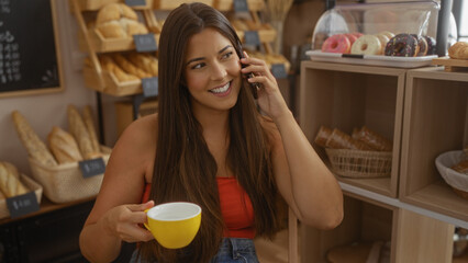 Young woman in a bakery talking on the phone and holding a yellow cup while smiling, surrounded by bread and pastries