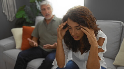 Woman stressed with man talking in background sitting on couch in living room showing middle age...