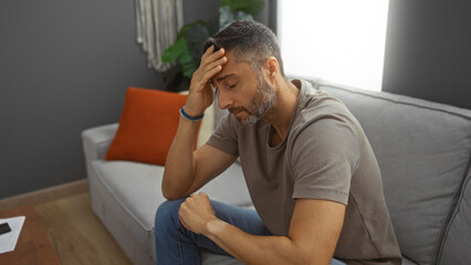 Young man looking worried sitting on a sofa in a cozy living room with hand on forehead, reflecting a sense of concern and introspection in a homely apartment setting.