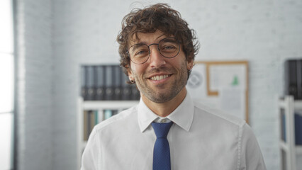 Handsome young man smiling in an office wearing glasses and a shirt with a tie, showcasing a...