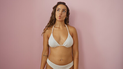Fototapeta premium Young woman posing in a white bikini over an isolated pink background, showing a playful expression with brunette hair and a beautiful, attractive appearance