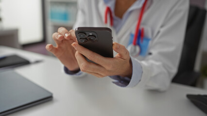 Woman in a clinic wearing a white coat uses smartphone at desk with focus on her hands and medical equipment visible, conveying theme of healthcare technology and communication.