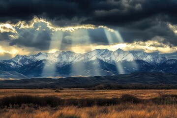 Sunlight breaks through dramatic clouds, casting radiant beams over a snowy mountain range and golden grassland below.
