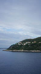Coastal view featuring lush hilltop houses under a cloudy sky beside calm sea reflecting a peaceful landscape.