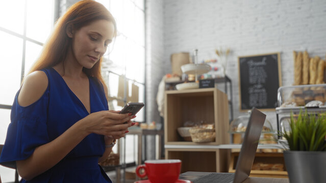 Woman in blue dress using smartphone in cozy bakery with laptop and red coffee cup on table, surrounded by shelves of bread and pastries.