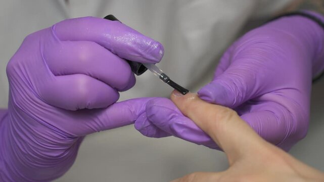 Professional manicurist wearing purple gloves applying clear nail polish to a client's fingernails