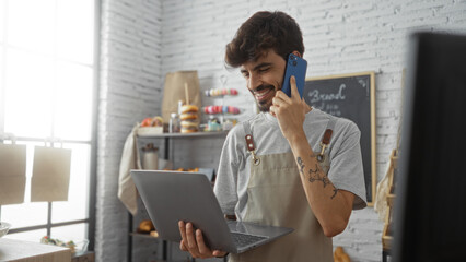 Young, handsome, hispanic man with a beard working in a bakery, smiling while talking on a smartphone and using a laptop in an indoor shop setting.