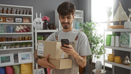 Handsome young man with a beard checking his mobile phone while holding packages in a decor store filled with various home decor items