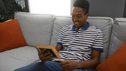 Young man smiling while sitting on a couch in a cozy living room holding a photo frame, showcasing a comfortable indoor environment filled with modern interior design elements.