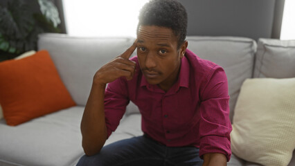 Young man in a living room touches his head thoughtfully against a backdrop of cozy cushions,...