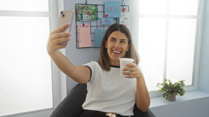 Young woman taking selfie in workplace holding coffee cup smiling in modern office interior with idea board in background
