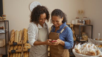 Two hispanic women bakers smiling and looking at a phone together in a bakery shop with various...