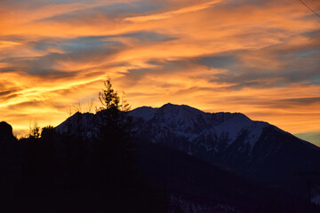 Golden hues of the sunset sky, blending vibrant shades of orange and yellow, cast a warm glow over the majestic High Tatras mountain peaks. Dark silhouettes of trees, dramatic contrast.