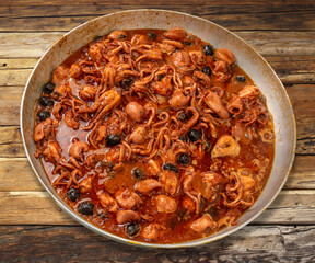 Baby Octopus in tomato sauce in aluminium pan on wooden table. Stewed octopus with tomatoes and olive - Italian recipe called moscardini alla Luciana recipe of Neapolitan cuisine.