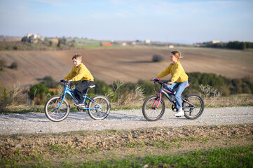 Obraz premium A boy and a girl race their bicycles through the countryside at sunset, laughing and chasing each other. The golden light and peaceful surroundings create a joyful, carefree moment of childhood advent