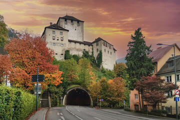 Medieval castle in Feldkirch, Austria, perched on a hill surrounded by vibrant autumn foliage, with a tunnel road under a dramatic sunset sky © runny1975