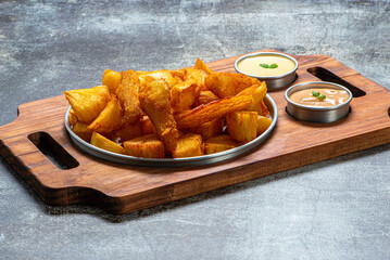 Porção de mandioca frita servida em tábua de madeira com molhos rosê e maionese
