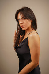 Fototapeta premium Attractive young woman with long straight hair and nude makeup in a sexy black dress in the studio. Charming lady posing for camera over gray background.