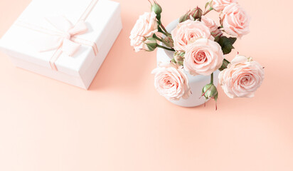 Bouquet of light roses on a beige table. Elegant bouquet of delicate peach roses in a white vase placed next to beautifully decorated gift on light surface, conveying love and celebration. 