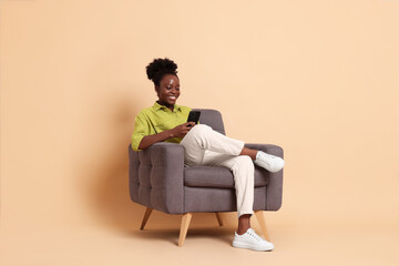 Smiling woman with smartphone sitting in armchair on beige background