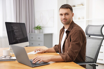 Programmer working on laptop and computer at wooden desk indoors