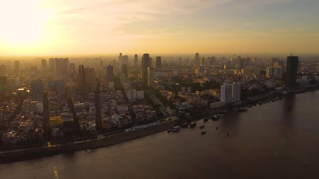 Drone view of densely populated Phnom Penh city at sunset with highrise commercial and residential buildings under hazy sky alongside Tonle sap river, Establishing shot