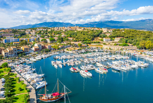 Aerial view of Porto-Vecchio marina in Corsica featuring luxury yachts, sailboats, and a scenic coastal village framed by mountains and the Mediterranean Sea.