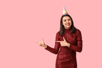 Beautiful young woman in party hat pointing at something on pink background