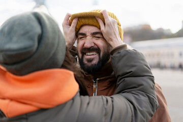 Woman pulling yellow beanie over man's head, making him grimace