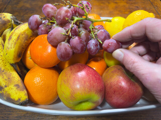 Closeup of a hand picking a grape from a juicy red bunch on top of a fruit bowl on a table, containing unpeeled, apples, oranges, bananas and lemons.