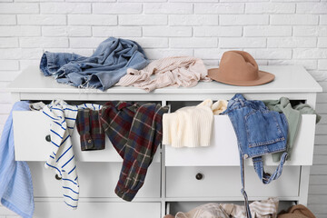 Stylish commode with different scattered clothes near white brick wall in room