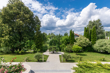 A picturesque landscape with flowering bushes and trees against a background of blue sky and white clouds. Park area with paths.