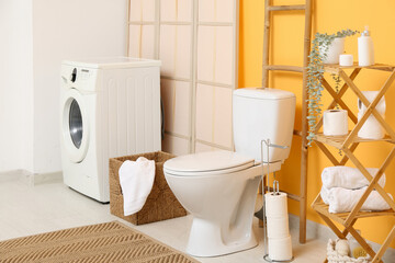 Interior of restroom with toilet bowl, washing machine and shelving unit near yellow wall