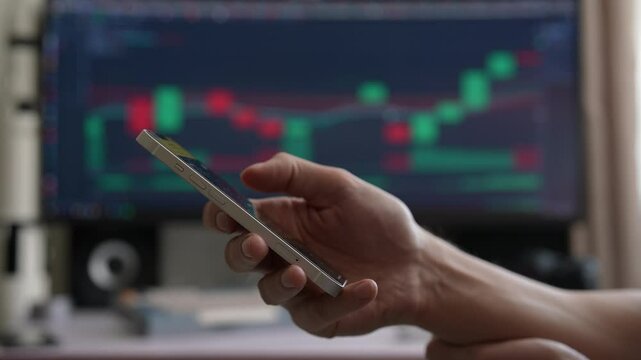 Side view of successful trader holding smartphone with trading app, while computer screen displays stock market charts, reflecting active market monitoring, closeup, unrecognizable person, slow motion