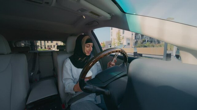 Pretty Arabic woman sitting in car holding coffee cup and smartphone. Female wearing hijab checking phone while parked near urban buildings. Casual morning routine or break outdoors.
