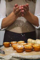 Close-up of hands sprinkling powdered sugar over freshly baked muffins on a wooden board, creating a cozy and appetizing baking scene.