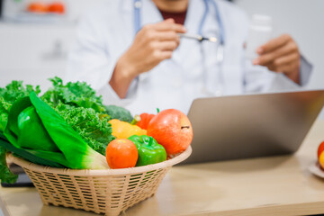 An Asian male nutritionist with a beard, wearing a lab coat, sits at a desk for an online consultation. His workspace includes fruits, vegetables, laptop, health supplements, promoting wellness.