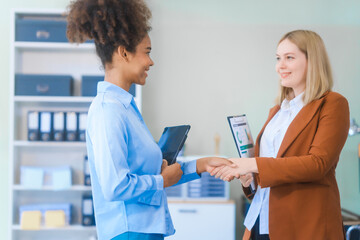 Obraz premium Two young professional women, an African American and a Russian, wearing formal suits, collaborate in a business office. They're consulting financial charts and graphs using desktop PCs, tablets