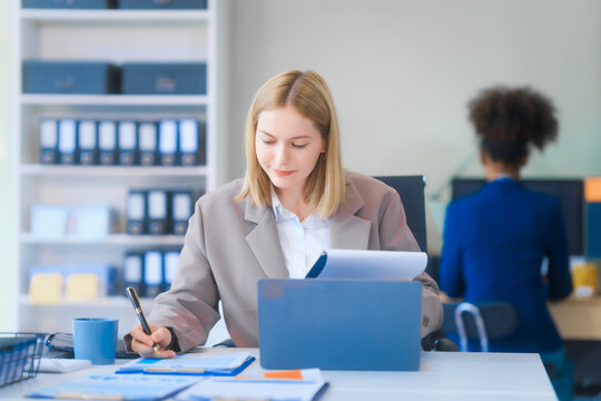 A young Russian woman in formal suit works diligently in business office, analyzing financial charts, using a desktop PC, calculator while contributing to various business strategies and projects.