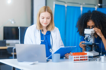 Young African American and Russian woman, both scientists, collaborate to analyze blood samples,...