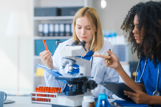 Young African American and Russian woman, both scientists, collaborate to analyze blood samples, viruses, vaccines using microscopes. They conduct crucial medical and biological research.