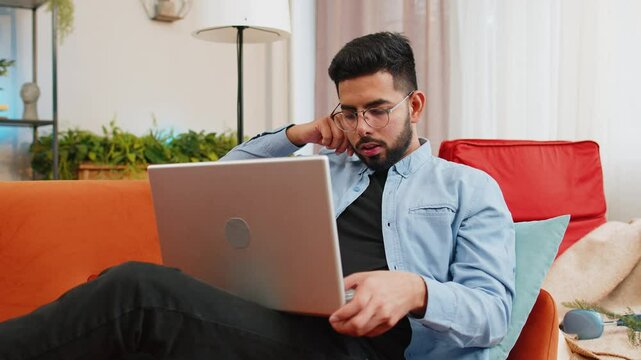 Close-up of surprised Indian man using laptop, receive good news shocked by sudden victory celebrate lottery jackpot win. Cheerful Hispanic guy with netbook sitting on sofa in living room at home.