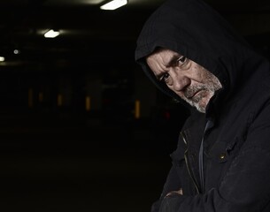 Portrait of an attractive older Caucasian man with grey hair and beard, wearing a hooded jacket. Serious expression. Dark, moody, industrial background.