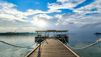 Obraz premium A serene wooden pier extends over calm water, leading to a small structure under a partly cloudy sky with the sun peeking through. The peaceful scene captures the tranquility of nature