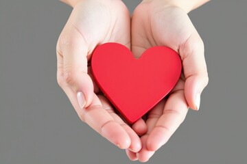 Obraz premium Close-Up of Hands Cradling a Vibrant Red Heart Symbolizing Love and Care Against a Neutral Gray Background