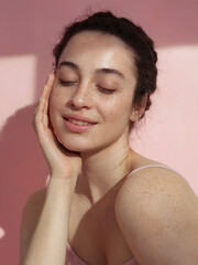 Naklejka premium self-love and self-confidence: A young woman with her eyes closed, exuding a serene expression. She has freckles on her face and is set against a soft pink background