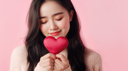A smiling Asian woman holding a red heart felt on a plain pink background with copy space for text.