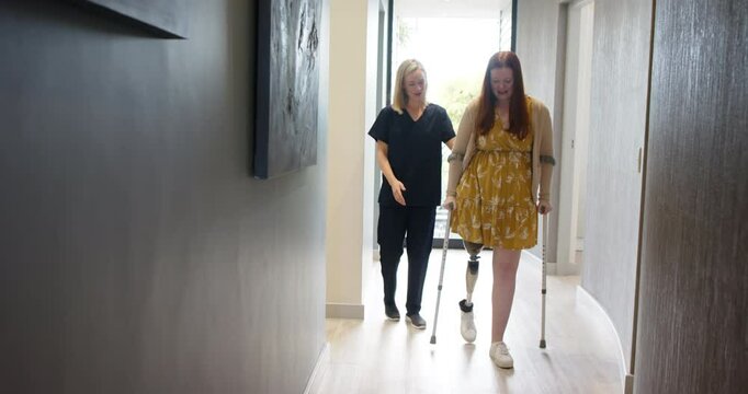 Woman with prosthetic leg walking using crutches with therapist in hallway, copy space