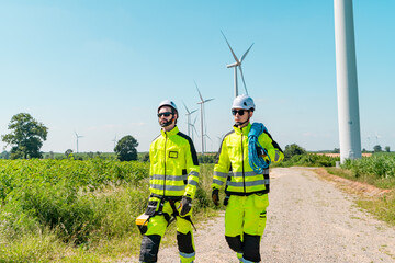 Two technicians walking along a pathway in a wind farm wearing safety gear during bright daylight